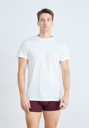 T-shirt en coton blanc à manches courtes, associé à un short boxeur ajusté bordeaux. Le mannequin se tient devant un fond clair, les bras détendus.