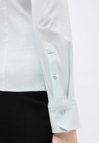 Λευκή μανσέτα από μακρυμάνικο πουκάμισο με δύο κουμπιά, φορεμένη με μαύρο κάτω μέρος, που δείχνει κοντινή εικόνα του μπράτσου και λεπτομέρειες της πλάγιας ραφής.