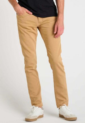 Man draagt een beige slimfit broek, met één hand in de zak, gecombineerd met witte sneakers en een zwart overhemd.