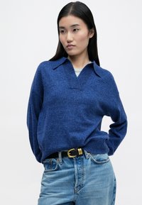 Blauer gestrickter Pullover mit Kragen, im V-Ausschnitt-Design und legerer Passform. Kombiniert mit hellblauen Jeans und einem schwarzen Gürtel.