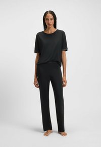 Vrouw die blootvoets staat, gekleed in een losse zwarte T-shirt met korte mouwen en bijpassende zwarte straight-leg broek, tegen een effen achtergrond.