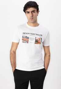 Camiseta de algodón blanca con estampados gráficos en naranja, gris y negro. Cuello redondo y mangas cortas, combinada con pantalones negros.
