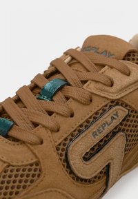 Καφέ σουέντ αθλητικό παπούτσι με πλέγμα, καφέ κορδόνια και μεταλλικές σμαραγδί οπές, με το λογότυπο Replay στο πλάι και στη γλώσσα.