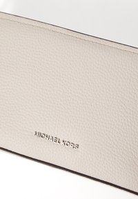 Beige läderplånbok med en strukturerad yta, prydd med en silvermetallisk logo "MICHAEL KORS" längst ner och kontrasterande sömnad i kanten.