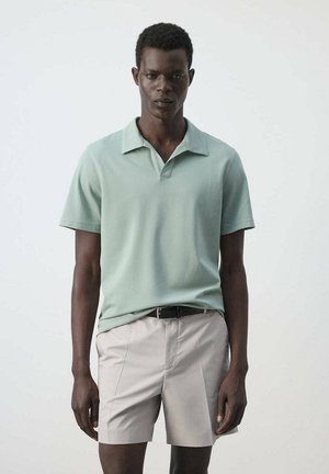Hombre con una camisa polo de manga corta verde claro y pantalones cortos beige con un cinturón negro, de pie frente a un fondo claro y liso.