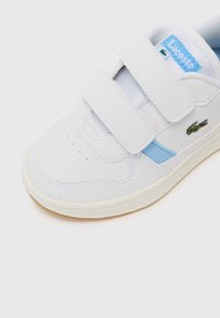 Baskets blanches avec des bandes Velcro, accent bleu, embout perforé pour la respirabilité et semelle en caoutchouc texturé. Logo Lacoste sur le côté.