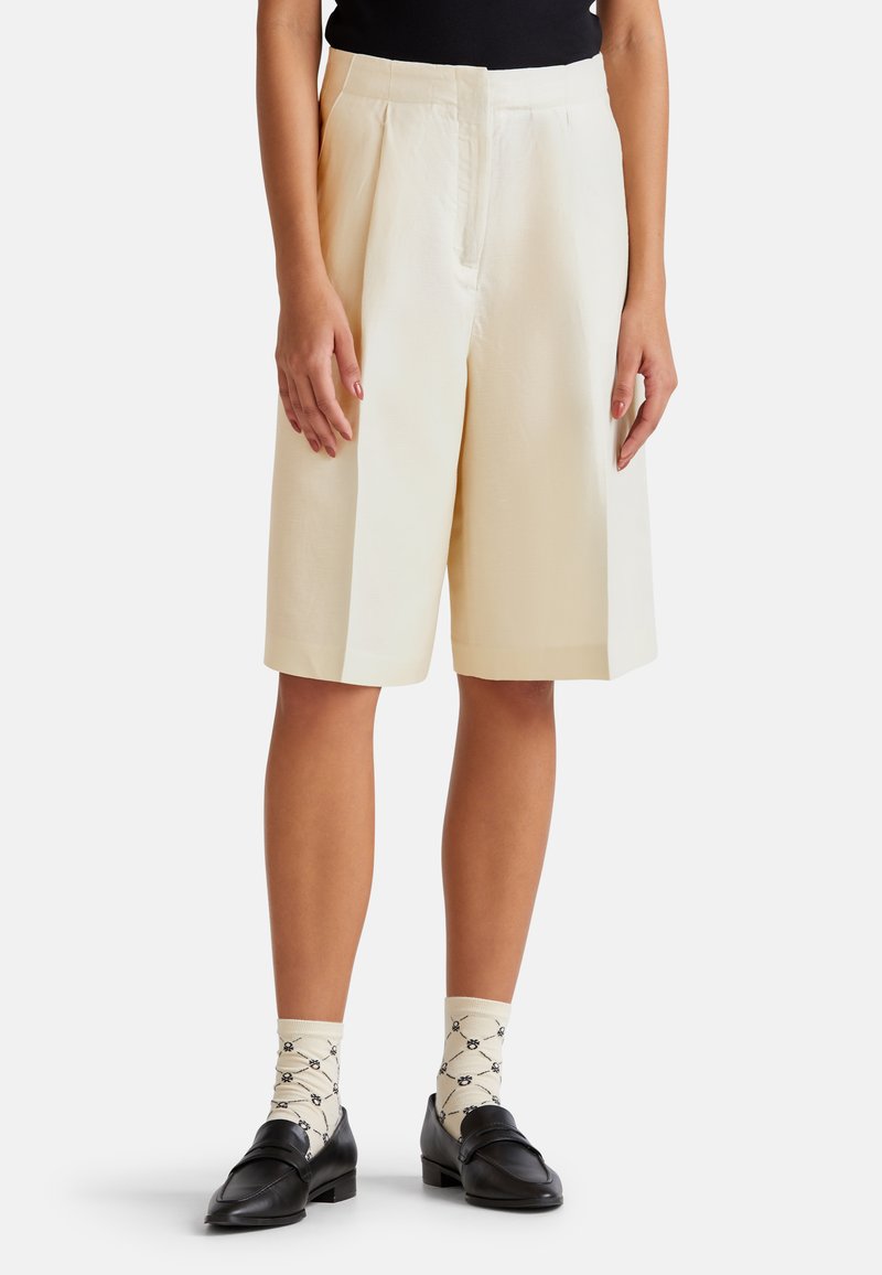 Ivory hoge taille shorts met plooien, losse pasvorm, gemaakt van een lichtgewicht stof. Draagt met gemusterde crèmekleurige sokken en zwarte loafers.