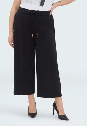 Kobieta ubrana w czarne szerokie spodnie 7/8 z troczkiem w pasie i czarne wysokie obcasy, stoi z jedną ręką na biodrze.