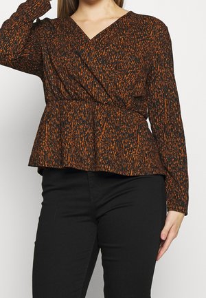 Vrouw die een blouse met lange mouwen en V-hals draagt met een zwart-oranje abstract patroon en zwarte aansluitende broek, poseert met één arm omhoog.