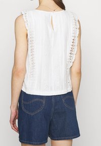 Vit ärmlös topp med spetskanter och vertikala ränder, med en nyckelhålsöppning bak, i kombination med mörka denimshorts.