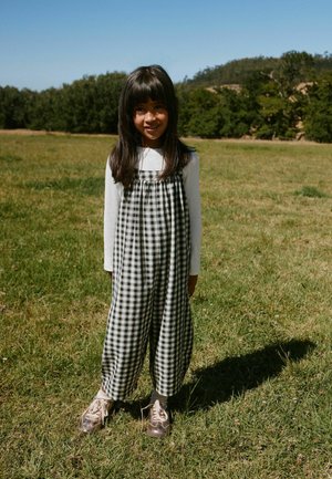 Junges Mädchen mit langen dunklen Haaren, das ein weißes Langarmshirt und schwarz-weiß karierten Overall trägt und auf Gras im Sonnenlicht steht.