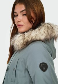 Parka grigio chiaro con colletto in pelliccia sintetica, chiusura con zip e tasche laterali. Presenta una patch con logo nera sulla manica.