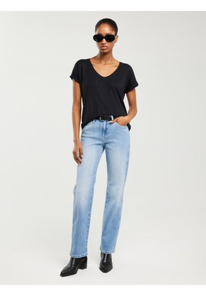 Nő fekete V-nyakú pólóban, világoskék farmerben, fekete övvel, fekete bokacsizmában és fekete napszemüvegben, egyszerű háttér előtt állva.