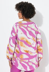 Maglietta a maniche lunghe di colore rosa con abstract bianchi e arancioni, caratterizzata da una vestibilità ampia e maniche a palloncino. La vista posteriore mostra l'orlo della maglietta.