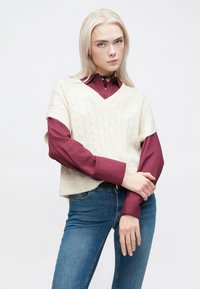 Ivory pletena telovnik s kabelskim vzorcem čez bordo dolgimi rokavi, z modrimi jeansi, ki prikazujejo sproščen kroj in plastenje.