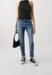 Svart stickad halterneck-top med vita prickar, ljusblå högmidjade jeans och svarta högstövlar, håller en liten svart handväska.