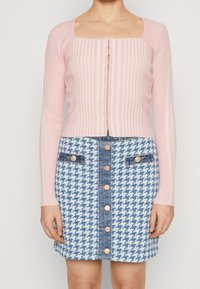 Top a maniche lunghe rigato rosa chiaro con scollatura quadrata e chiusura a bottoni, abbinato a una gonna di jeans con motivo a scacchi e dettagli metallici.