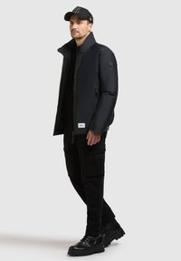 Mężczyzna w czarnej pikowanej kurtce, czarnych spodniach cargo, czarnych butach i czapce z daszkiem stoi na szarym tle.