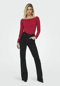 Crvena bluzica dugih rukava s širokim dekolteom uparena s crnim trapericama visokog struka i širokim nogavicama. Model ima ruke u džepovima, nosi crne cipele s oštrim vrhom.