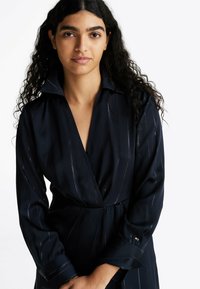 Vestido tipo camisa azul marino con cuello, con rayas verticales brillantes, mangas largas y cintura con cinturón. Textura suave, diseño elegante.