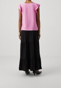 Kvinna som står med ryggen mot kameran, iklädd en rosa topp med volanger, lång svart flerlagerskjol och svarta kitten heal-skor, på vit bakgrund.