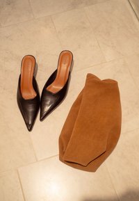 Een paar zwarte pumps met puntige neus naast een opgevouwen bruine suède jas op een beige betegelde vloer.