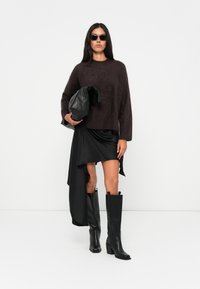 Pullover marrone peloso, gonna asimmetrica in satin nero, alti stivali in pelle nera e una borsa a tracolla nera oversized. La modella indossa occhiali da sole scuri.