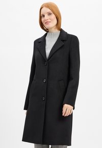 Eine rothaarige Frau trägt einen schwarzen Knopfmantel über einem hellgrauen Pullover vor einem schlichten weißen Hintergrund.