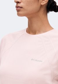 Γυναίκα με ανοιχτό ροζ μακρυμάνικη μπλούζα Columbia, προφίλ από το πλάι που δείχνει τον λαιμό, τη γραμμή της γνάθου και το άνω μέρος του στήθους σε μονόχρωμο φόντο.