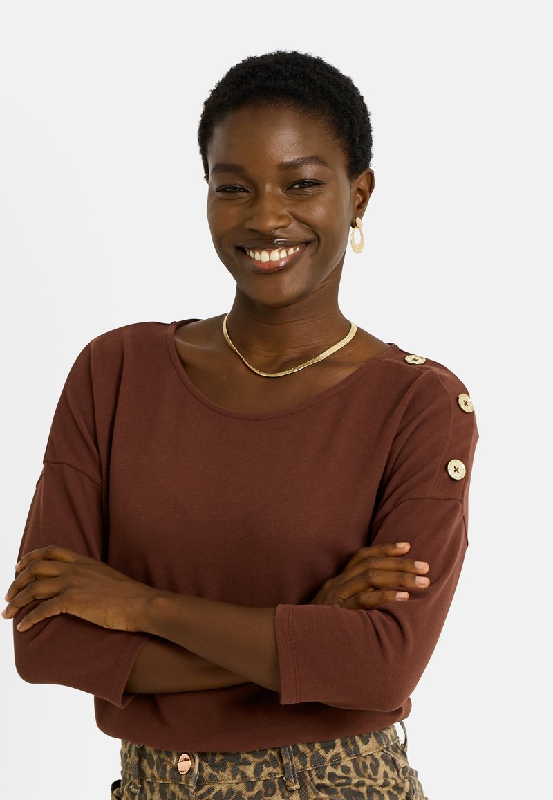 Top marrón de manga larga con cuello redondo, con botones decorativos en los hombros, combinado con pantalones con estampado. Textura lisa.