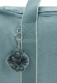 Lichtblauwe tas van stof met een rits, met een fuzzy grijze gorilla sleutelhanger bevestigd aan een metalen ring aan de zijkant.