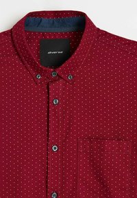 Bordowa bawełniana koszula w białe kropki, z kołnierzykiem na guziki, kieszenią na piersi oraz kontrastowym granatowym wewnętrznym kołnierzem. Posiada niebieskie guziki.