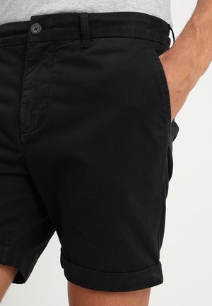 Mann trägt schwarze Shorts mit aufgerolltem Saum, vorderem Knopf und Seitentaschen, eine Hand teilweise in der Tasche, vor neutralem Hintergrund.