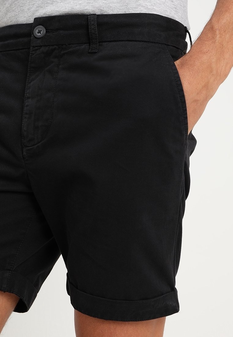 Homme portant un short noir à ourlet roulé avec un bouton à l'avant et des poches latérales, une main partiellement dans une poche, sur un fond neutre.