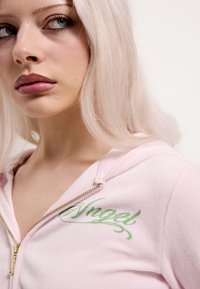 Różowa bluza na suwak z zielonym haftowanym napisem „Angel” na lewej piersi, o gładkiej fakturze i złotym zamku błyskawicznym.