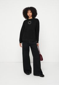 Μαύρο φούτερ με στρογγυλό λογότυπο στο κέντρο, συνδυασμένο με μαύρο τζιν παντελόνι με φαρδιά πόδια και μικρή μπορντό τσάντα.