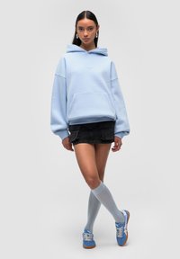 Hoodie overdimensionato azzurro con tasca frontale, abbinato a una mini gonna nera. Sneakers blu e calze grigie al ginocchio completano l'outfit.