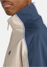 Jacket featuring a high collar with a two-tone design: soft beige fabric on one side and dark blue on the other, accented with light stitching.