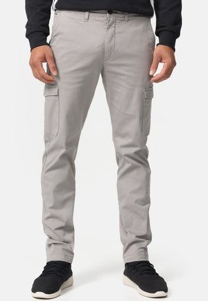 Hombre con pantalones cargo ajustados de color gris claro con bolsillos laterales con solapa y zapatillas negras, de pie sobre un fondo blanco.