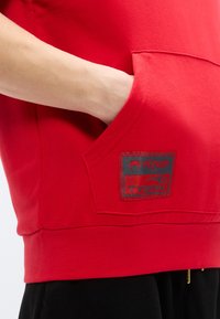 Κόκκινο βαμβακερό φλις με τσέπη καγκουρό και μαύρο κονκάρδα με τα λογότυπα "VENUM" και "TYSON". Λεία υφή και άνετη εφαρμογή.