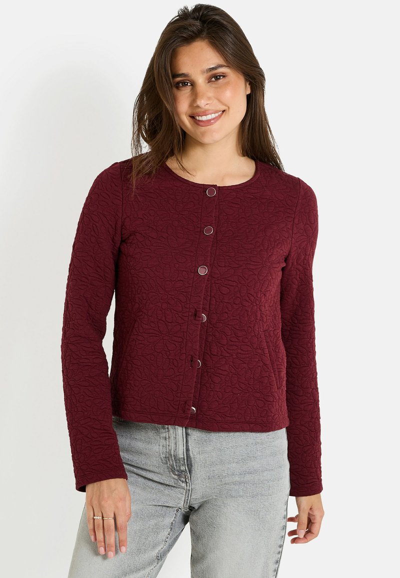 Femme souriante portant une veste boutonnée en maroon texturé et un jean gris clair, se tenant avec une main détendue à ses côtés.