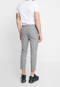 Muž v šedých kostkovaných slim-fit zkrácených kalhotách, bílém tričku a černých teniskách, stojí zády k fotoaparátu.