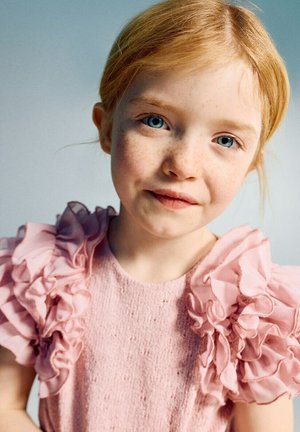 Ragazza giovane con capelli rossi e lentiggini che indossa un vestito rosa con texture e maniche arricciate, che guarda direttamente la fotocamera con un leggero sorriso.