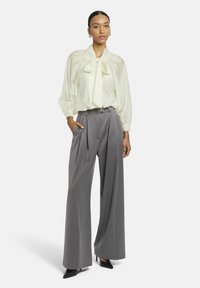 Cremefarbene Bluse mit Schleife und Puffärmeln, kombiniert mit hoch taillierter, grauer Plisseehose. Das Model trägt schwarze spitze Pumps.