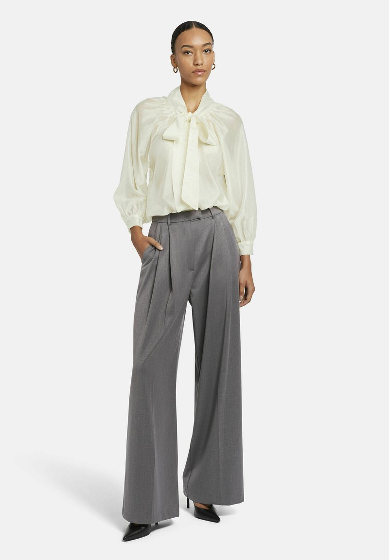 Cremefarbene Bluse mit Schleife und Puffärmeln, kombiniert mit hoch taillierter, grauer Plisseehose. Das Model trägt schwarze spitze Pumps.