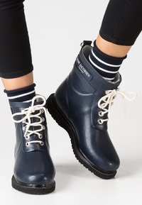 Botas de goma azul marino hasta el tobillo con cordones blancos y una etiqueta con el logo. Presentan una textura suave y puntera redonda, complementadas con calcetines a rayas.