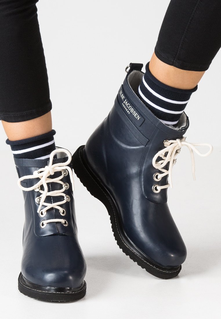 Botas de goma azul marino hasta el tobillo con cordones blancos y una etiqueta con el logo. Presentan una textura suave y puntera redonda, complementadas con calcetines a rayas.