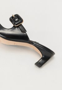 Musta nahkasandaali matalalla lohkoltaalla, jossa on neliömäinen solki ja avonainen varvasdesign. Sileä pinta ja neutraali sisävuori.