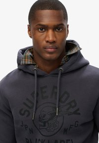 Marine hoodie met een grafisch schedelontwerp, voorzien van een geruite voering in tinten groen en oranje, trekkoorden en metalen accenten.