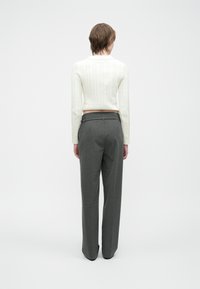 Person, der mit dem Rücken zur Kamera steht und einen weißen Kabelstrickpullover sowie graue elegante Hosen trägt, vor einem schlichten Hintergrund.